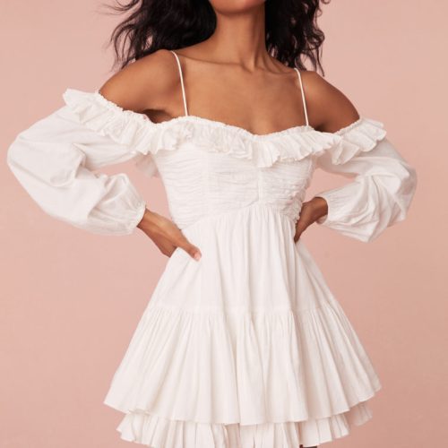 White mini dress featuring a shaped waist with shirring and ruching at the bust. Includes off-shoulder blouson sleeves, elasticated openings, a two-tiered skirt, a smocked back waist, and adjustable spaghetti straps.